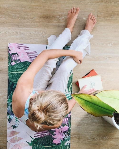 A close-up shot of a hands performing a yoga pose on a colorful yoga mat. The mat is placed on a hardwood floor, and sunlight streams in from a nearby window.