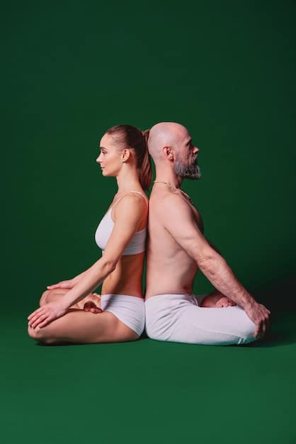 Two people doing a seated twist in partner yoga, facing each other and holding hands, with a focus on their spinal alignment and synchronized breathing in a serene studio setting.