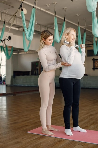 A yoga instructor demonstrating a modified warrior pose to a pregnant woman in a studio setting. The instructor is showing how to adjust the stance to accommodate the woman's growing belly.