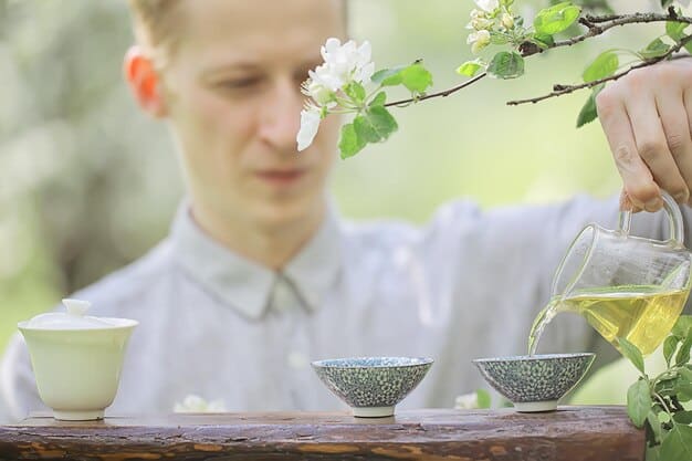 A person pouring tea into a small teacup with focused attention, with a blurred background of a peaceful garden.