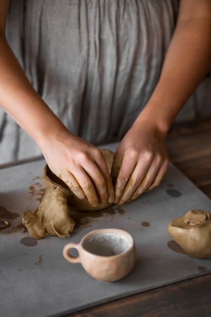 A close-up shot of an artist's hands gently shaping clay on a pottery wheel. Soft, diffused light highlights the texture of the clay and the artist's focused expression.