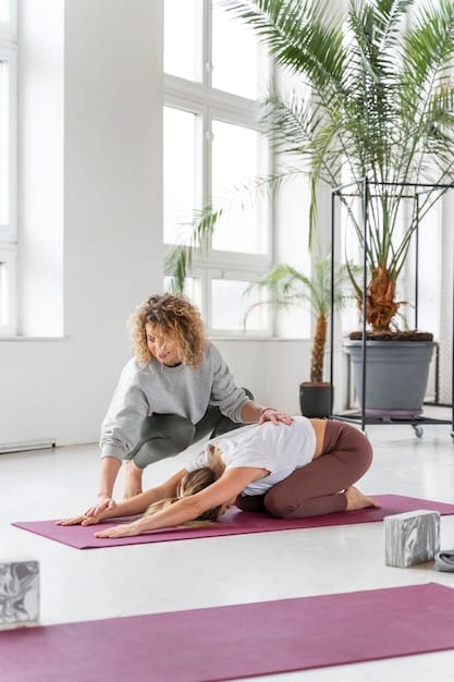 A yoga instructor assisting a student in a seated forward fold pose. The instructor is gently guiding the student's back, encouraging a deeper stretch while ensuring proper form. The student is seated on a yoga mat with legs extended, reaching towards their toes.