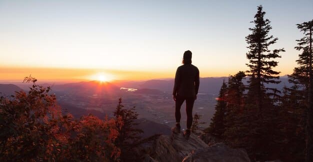 A person standing on a mountaintop, looking out at a vast, scenic landscape. The sun is setting, casting a warm glow over the hills and valleys. The image conveys a sense of accomplishment, peace, and connection with nature.