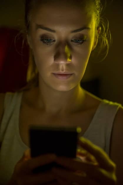 A close-up shot of a person's anxious face illuminated by the glow of a smartphone screen in a dark room. The focus is on the tension in their brow and the distracted look in their eyes.