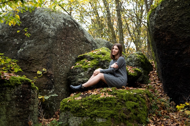 A person sitting in a meditative pose on a moss-covered rock in a lush forest. Sunlight filters through the trees, highlighting the individual's serene expression and relaxed posture. The background includes a small waterfall and various shades of green, creating a sense of harmony with nature.