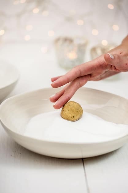 A close-up shot of a hand gently pouring water into a bowl in a Zen garden. The water is clear, and the bowl is simple and rustic. The background shows raked sand and a few carefully placed rocks.