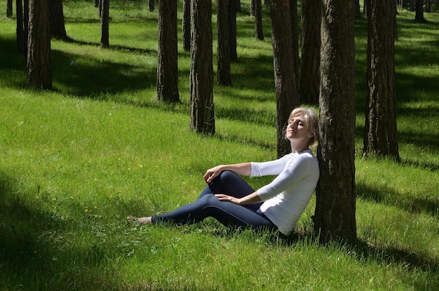 A person sitting in a comfortable position, eyes closed, meditating outdoors in a natural setting. Sunlight filters through the trees, creating a serene atmosphere.